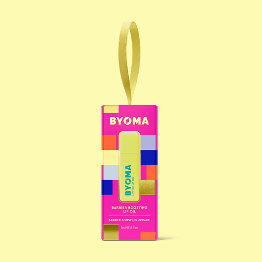 BYOMA Barrier Boosting Lip Oil in pink and gold gift box with hanging ribbon, displayed on yellow background