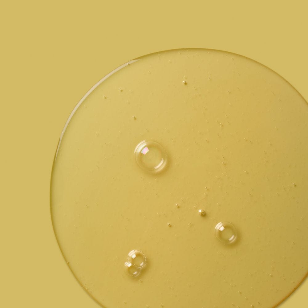 Macro close-up of translucent yellow serum or oil with several suspended bubbles, placed against a matching yellow background.
