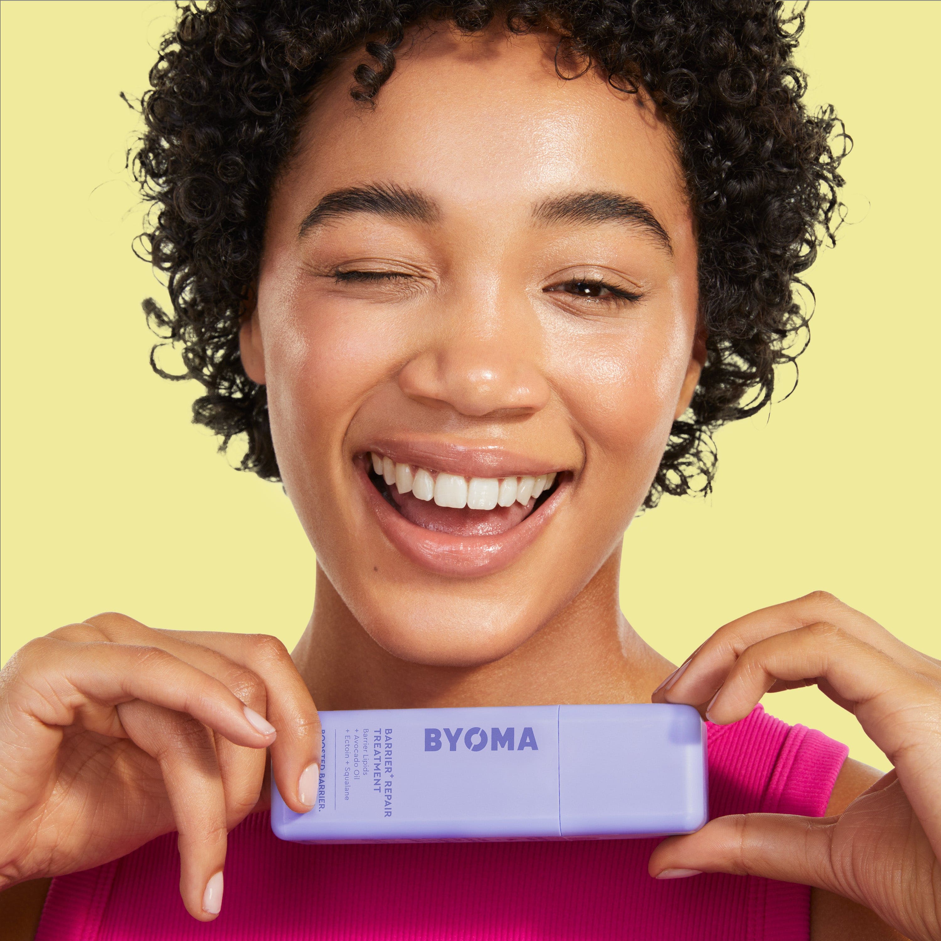 A winking and smiling girl horizontally holding a bottle of BYOMA BARRIER REPAIR TREATMENT.