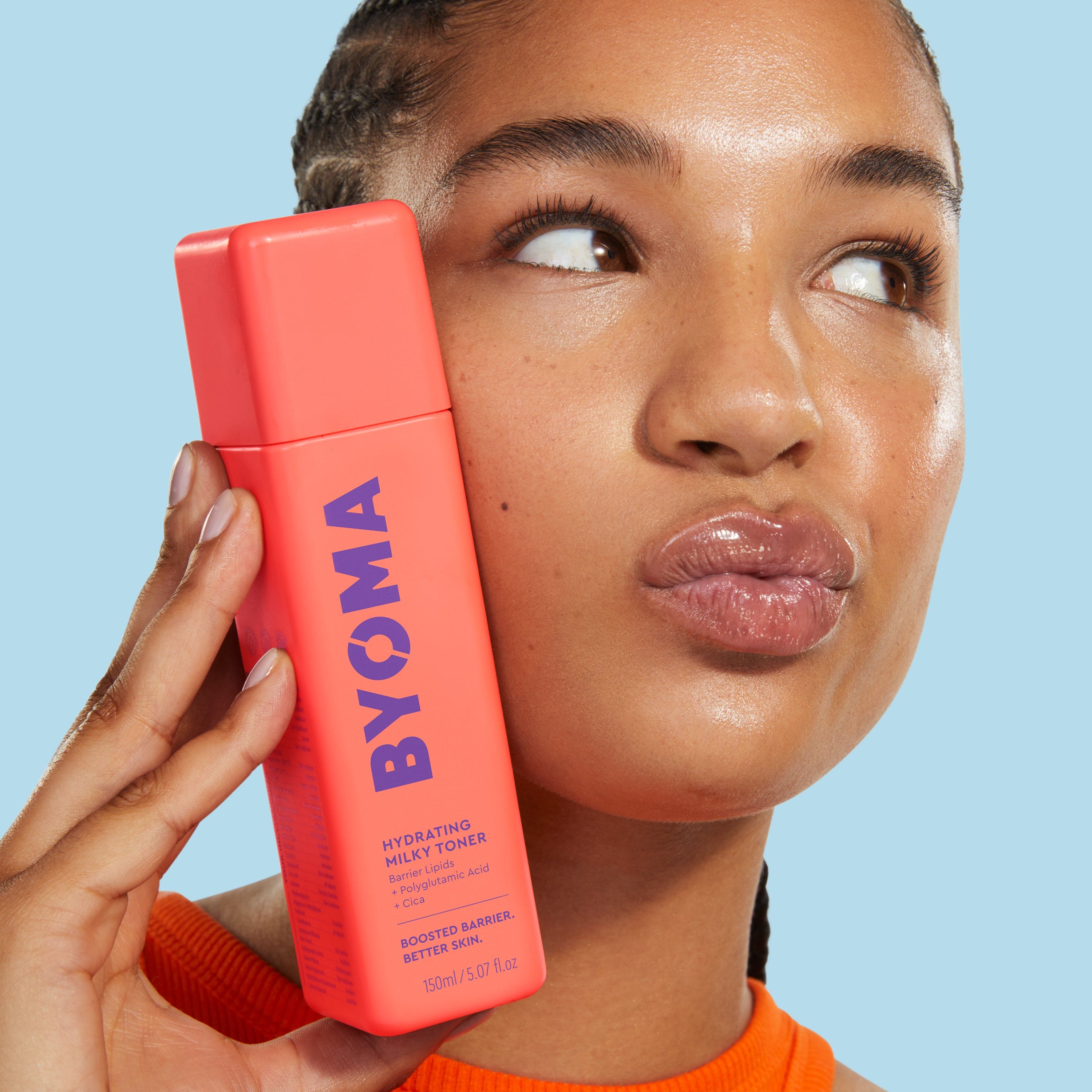 A girl in orange tank top with puckered lips holding a bottle of BYOMA product against her face.