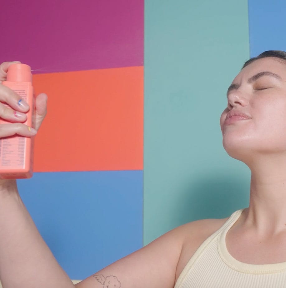 A woman with closed eyes misting a BYOMA product on her face and a colourful background.